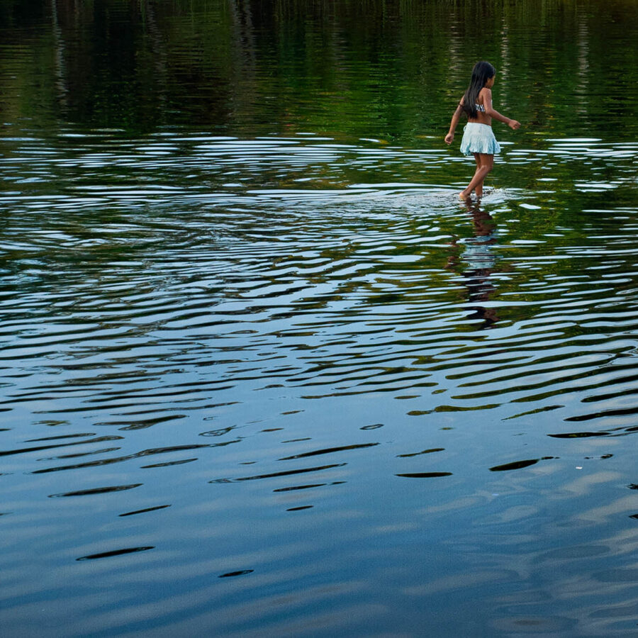 Niña pequeña vestida de blanco caminando sobre la superficie de un cuerpo de agua, con su reflejo visible debajo. El agua presenta suaves ondas.