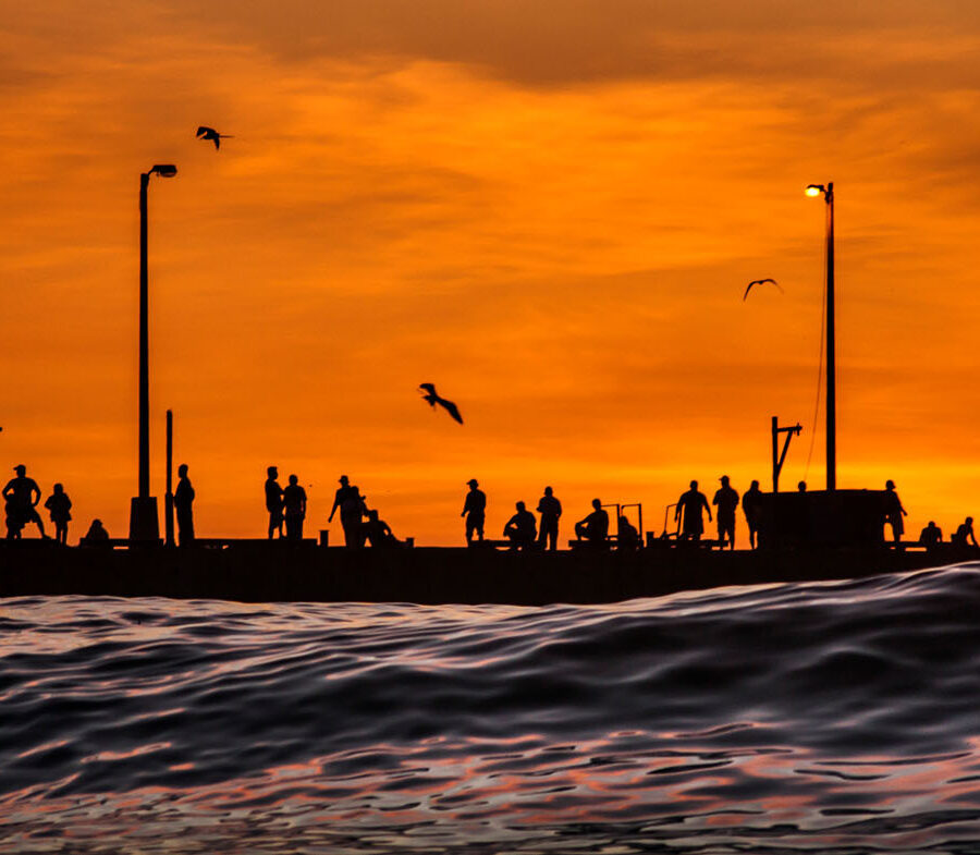 Silueta panorámica del Muelle de Pimentel en la ciudad de Chiclayo, Perú. Varias personas de pie, recortadas contra un cielo de atardecer de color naranja intenso y vibrante sobre el mar.