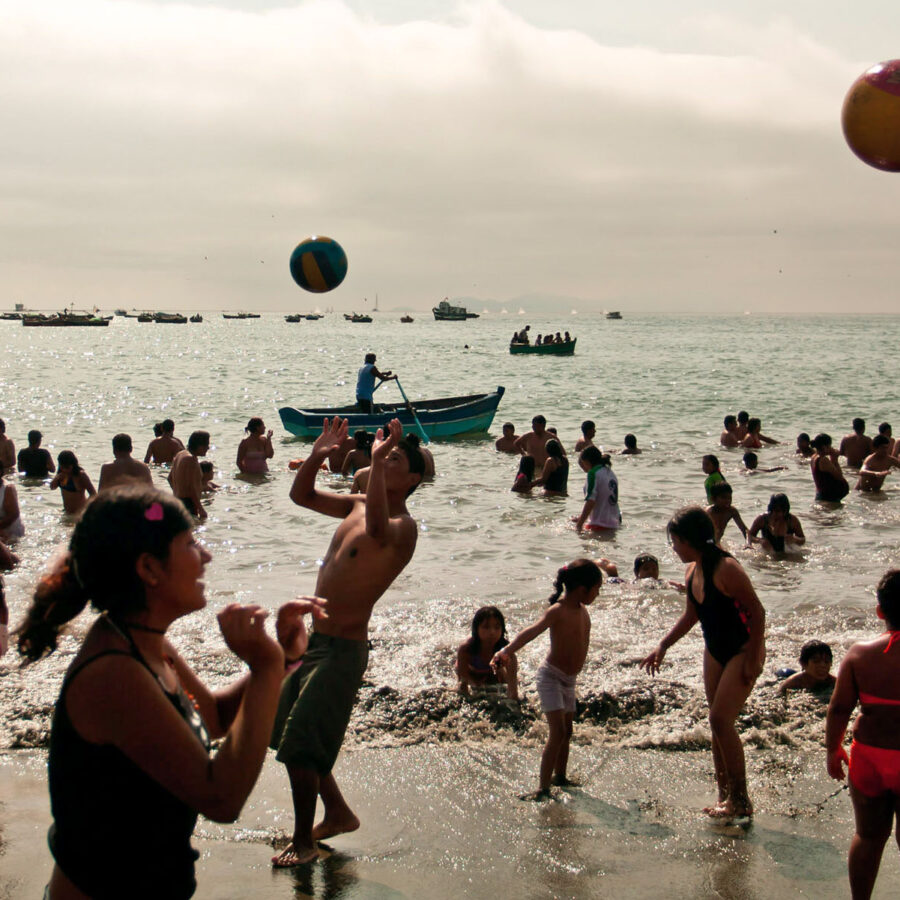 Grupo de personas jugando con una pelota grande en la orilla de la playa Aguadulce en un día nublado, con botes de pesca visibles en el horizonte.