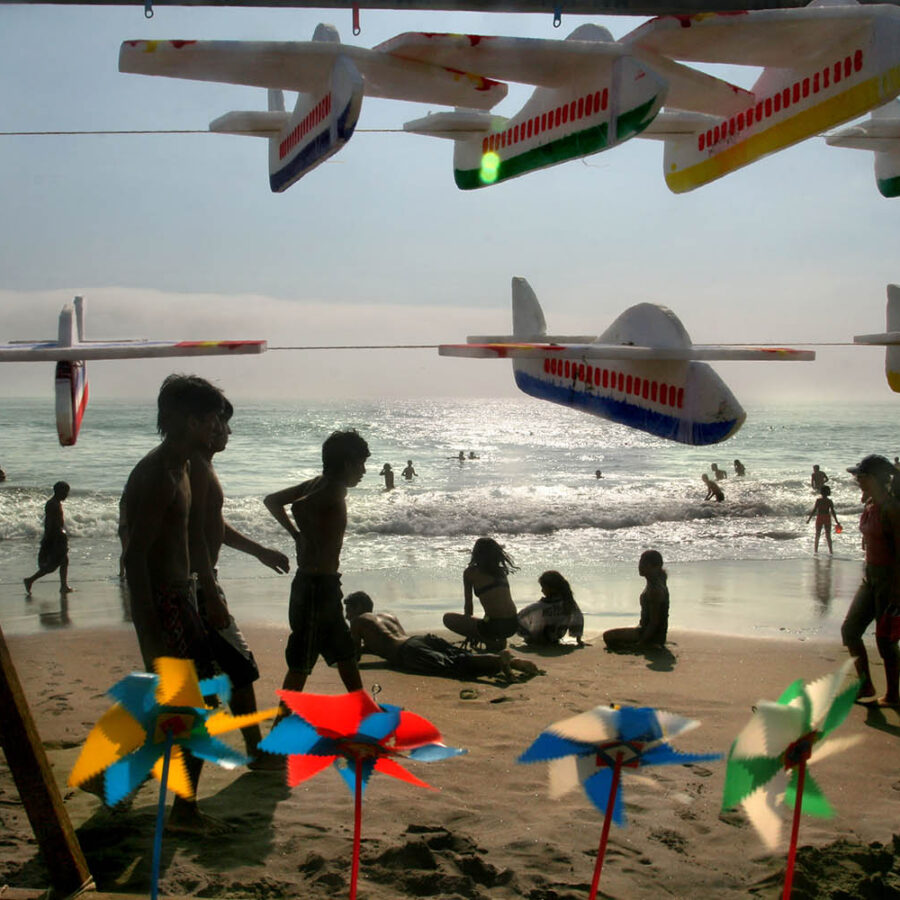 Vista de la playa Aguadulce a través de juguetes de espuma (aviones) y molinillos de viento colgados en primer plano, con siluetas de personas al fondo.