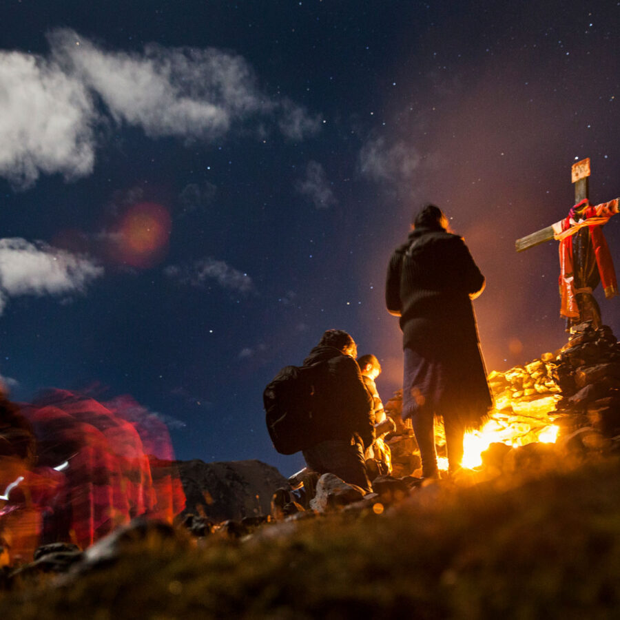 Escena nocturna de la celebración de Qoyllur Riti, con devotos reunidos alrededor de una cruz adornada e iluminada por una fogata bajo un cielo estrellado.