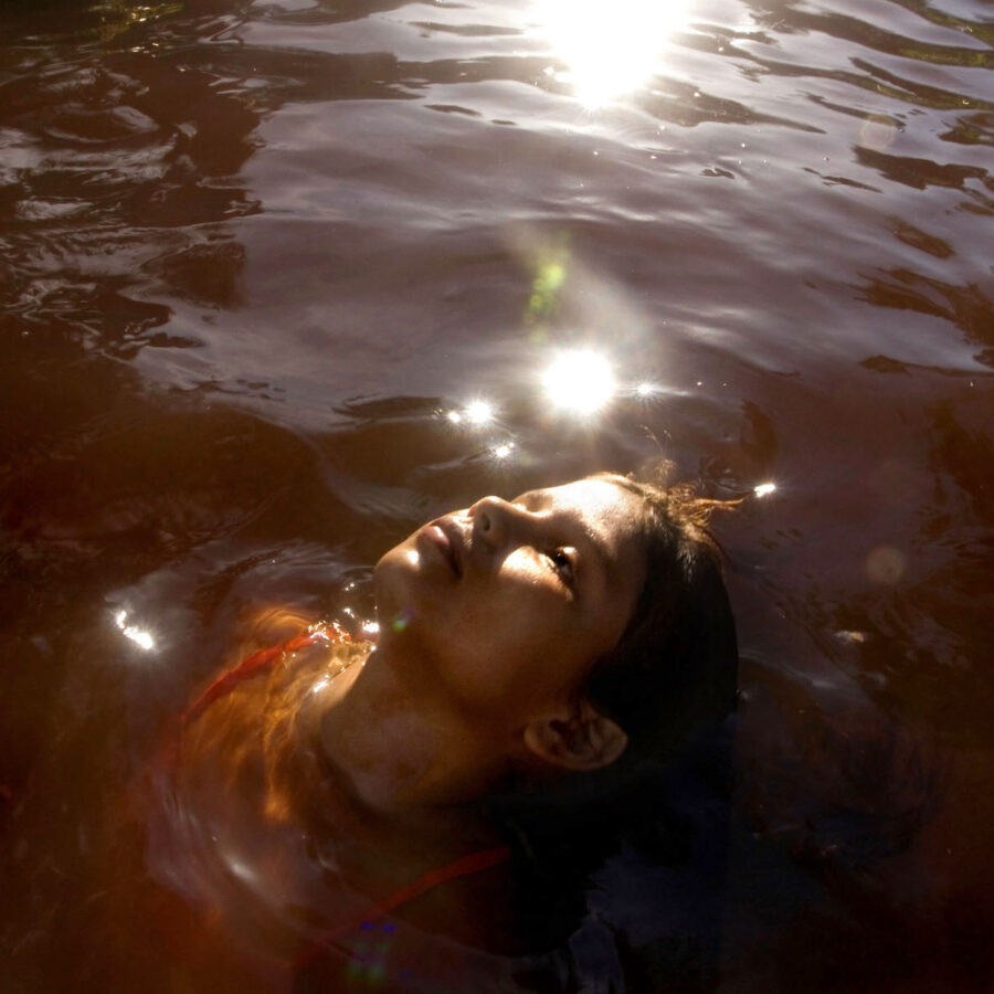 Retrato cenital de una persona flotando relajadamente boca arriba en las aguas rojizas del río Corrientillo, con destellos de luz solar reflejándose en la superficie.