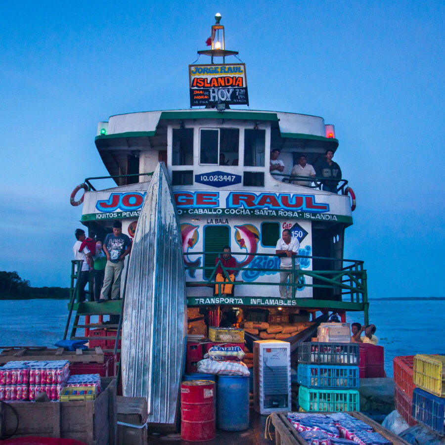 El barco fluvial de carga y pasajeros llamado "Jorge Raul" navegando por el río Amazonas al atardecer, cargado con cajas de mercancía y gente en cubierta.
