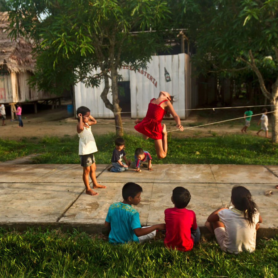 Un grupo de niños jugando a saltar la liga al aire libre en la comunidad de Pucaurquillo, Perú. Una niña salta en el aire y otros niños observan sentados.