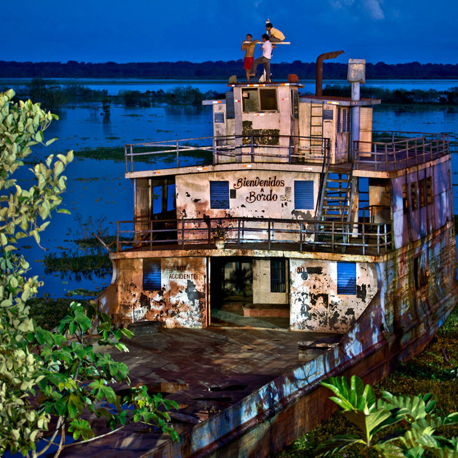 Barco oxidado y antiguo varado entre vegetación al atardecer en el río Itaya, con la inscripción visible "Bienvenidos a bordo" en la fachada. Desde él tres amigos contemplan el paisaje.