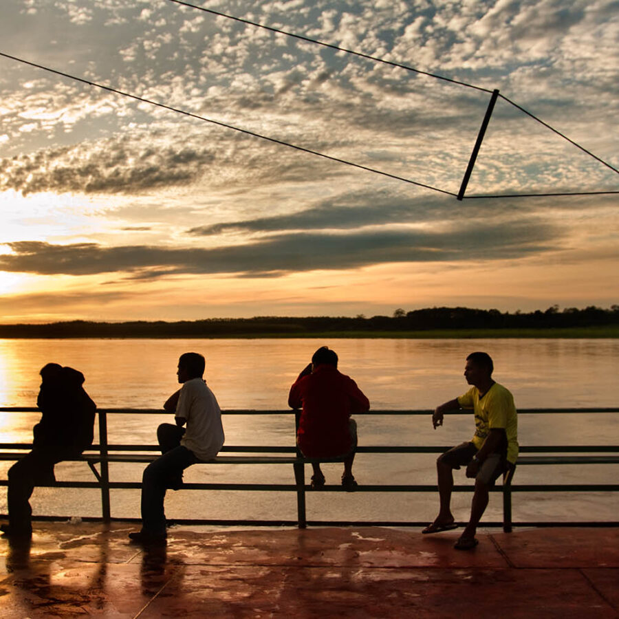 Siluetas de un grupo de personas sentadas en la baranda de una embarcación fluvial navegando por el río Amazonas, bajo un cielo de atardecer con nubes texturizadas.