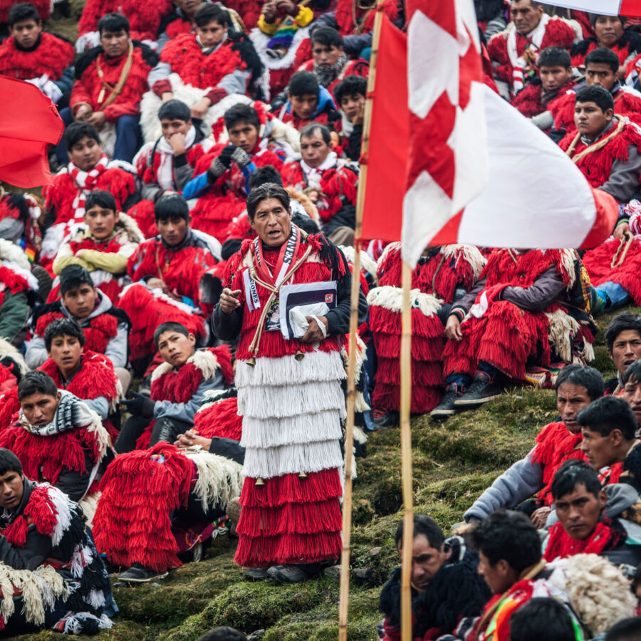 Multitudinaria comparsas de la nación Quispicanchis en asamblea comunitaria en las celebraciones del Señor de Qoyllur Riti. Asistentes vistien ponchos rojos tradicionales y banderas peruanas, escuchando a un orador en el centro.