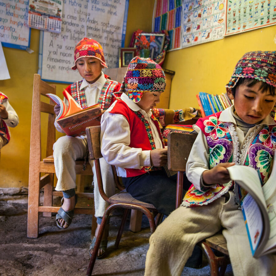 Niños estudiantes vistiendo chullos y chalecos tradicionales bordados, leyendo concentrados en sus pupitres dentro de una escuela rural en Pitumarca. Canchis, Cusco, Perú