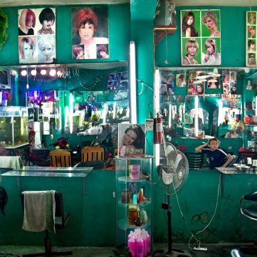 Interior de una barbería o salón de belleza con paredes verde azulado brillante. Hay espejos, luces de neón, sillas de peluquería y recortes de revistas con peinados en las paredes.