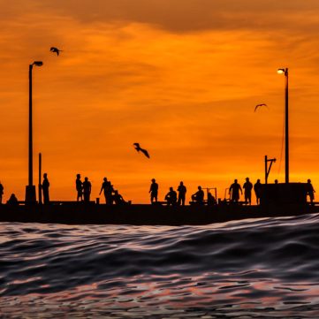 Silueta panorámica del Muelle de Pimentel en la ciudad de Chiclayo, Perú. Varias personas de pie, recortadas contra un cielo de atardecer de color naranja intenso y vibrante sobre el mar.