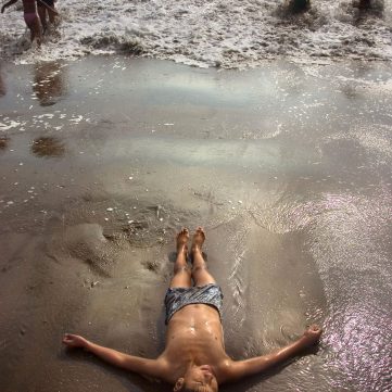 Niño tumbado boca arriba con los brazos abiertos en la orilla mojada de la playa Aguadulce, con bañistas disfrutando de las olas al fondo.