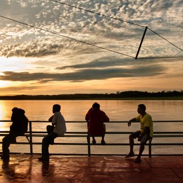 Siluetas de un grupo de personas sentadas en la baranda de una embarcación fluvial navegando por el río Amazonas, bajo un cielo de atardecer con nubes texturizadas.