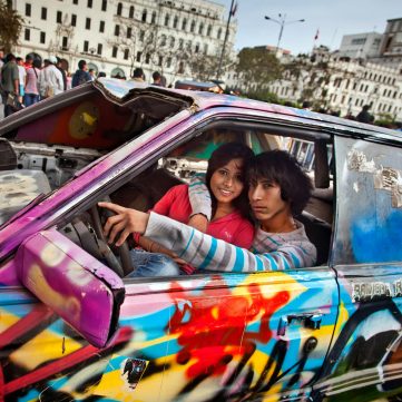 Retrato de una pareja joven en un coche pintado con graffiti, rodeados de gente, en la plaza San Martín de Lima, Perú.